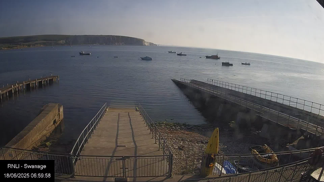 A view of a harbor with a concrete pier extending into calm water. Several boats are anchored in the distance, and a larger landmass with cliffs and greenery is visible on the horizon. The sky appears clear with soft lighting, suggesting early morning. A yellow buoy and some boating equipment are near the foreground. The timestamp indicates the image was captured on June 18, 2025, at 6:59 AM, and the location is Swanage.