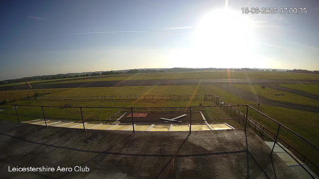 The image shows a bright, sunny day at Leicestershire Aero Club with clear blue skies and some faint contrails. The scene is captured from a balcony or observation area overlooking a grassy field and a track or runway in the background. The sun is high and shining strongly, creating lens flare and long shadows. The timestamp in the top right corner reads "18-06-2025 07:00:35". The bottom left corner has the text "Leicestershire Aero Club."
