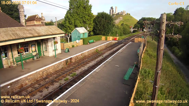 A small train station platform with a stone building featuring green benches, hanging flower baskets, and a small green shed nearby. The railway tracks run through the scene, with a grassy hillside and a castle ruins visible in the background on a sunny day. Two utility poles with wires are on the right side, and there are several benches along the platform. The scene appears peaceful with lush green trees and a clear sky.