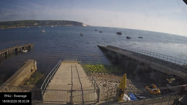 A coastal scene with a small harbor. There is a concrete pier extending into the water, with a ramp leading down to the shore. Several small boats are moored near the pier, and a few are in the distance on the water. A yellow signboard and some equipment are visible on the shore. In the background, there are green hills with a few white cliffs along the coast. The sky is partly cloudy, and the time shown is 8:00 AM.