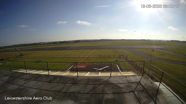 A view from a webcam at Leicestershire Aero Club shows a clear, sunny day with a few scattered clouds. In the foreground, there is a fenced area with a small aircraft lying on its side, suggesting it may be under maintenance or storage. The background displays a runway extending into a grassy field, with a distant horizon of trees and open land. The timestamp on the image indicates it was taken on June 18, 2025, at 8:00 am.