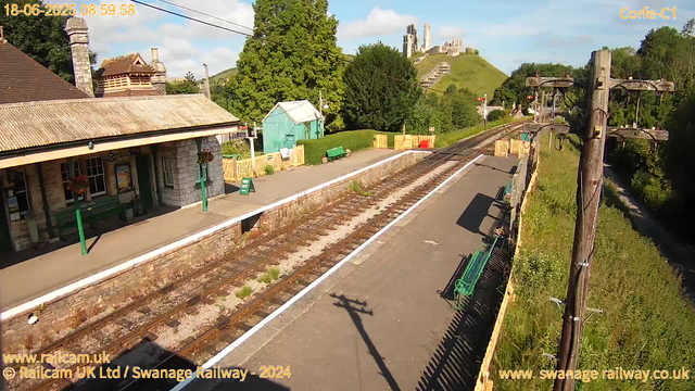 A small rural train station platform with a building on the left, featuring a chimney and window boxes with flowers. The platform has two green benches, a sign reading "Way Out," and some potted plants. Overhead, there are wires for the train. The railway tracks run through the center, with a fence separating the platform from the surrounding greenery. In the background, lush trees and a hill with ancient castle ruins on top are visible under a partly cloudy sky. The timestamp in the top left corner reads "18-06-2025 08:59:58," and the top right corner shows "Corfe-C1."