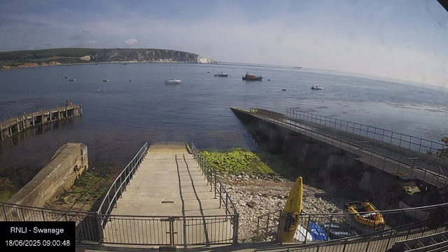 A calm seaside scene with a concrete pier extending into the water, two yellow boats parked on the shore, and several boats anchored further out. In the background, there are white chalk cliffs and a hilly landscape under a partly cloudy sky.
