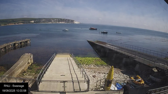 A view of a harbor with calm water, several boats and ships anchored in the distance, a cliff with white chalk faces on the horizon, and a partly cloudy sky overhead. In the foreground, there is a small concrete pier with metal railings, a yellow life raft, and a person standing near the railing.