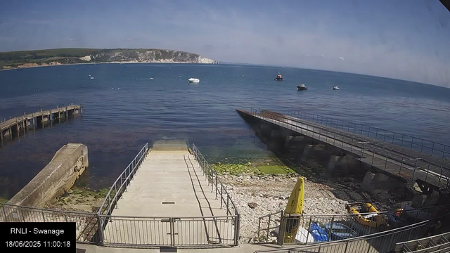 A calm seaside scene with a small dock extending into the water, surrounded by railings. The shoreline has rocks and some green algae or seaweed. Several boats are anchored offshore in the water, with a cliff and green landscape in the background under a partly cloudy sky. Near the dock, there is a yellow caution sign, a small yellow boat, and some equipment or supplies.
