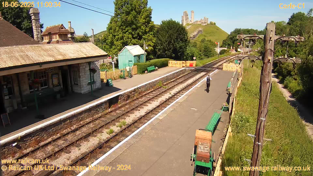 A small rural train station platform with a single person walking away from the camera. There are green benches along the platform, with a wooden fence and greenery behind it. A utility pole with wires runs alongside the platform. The station building is on the left, made of stone with a tiled roof. In the distance, a hill with the ruins of a castle is visible under a clear blue sky. The timestamp on the image reads "18-06-2025 11:00:34".
