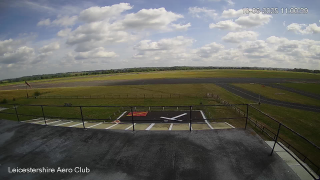 A view from a webcam at Leicestershire Aero Club showing an open grassy field with a small helicopter landing pad in the foreground. The sky is partly cloudy with patches of blue, and there are distant fields and trees on the horizon. The date and time are displayed in the upper right corner as 18-06-2025 11:00:29.