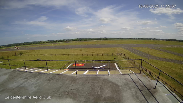 A view from a webcam at Leicester Aeroclub shows a clear sky with some scattered clouds. In the foreground, there is a small parking area with painted markings and a windsock on a pole. Beyond the parking area, there is a grassy apron and a paved taxiway leading to a runway in the distance. The landscape is flat with fields and distant trees on the horizon. The image is timestamped "18-06-2025 11:59:57".