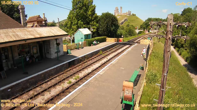 A small train station platform with a stone building on the left, a few people sitting on benches, and some greenery in the background. Railway tracks run through the center, and a hill with a castle on top is visible in the distance. The sky is clear and blue, and there are a few utility poles and overhead wires along the platform.