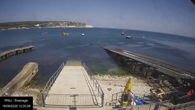 A view of a coastal area with a small dock, several boats on the water, and a landmass with cliffs and green hills in the background. The scene is bright and clear, with a blue sky overhead. In the foreground, there are people near the dock, some with yellow inflatable boats and equipment. The timestamp shows the date as June 18, 2025, at 12:00:58, and the location is identified as RNLI - Swanage.