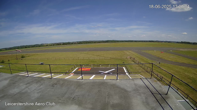 A view from a webcam at Leicesterhire Aero Club shows a clear day with a blue sky and some scattered clouds. The foreground features a helipad with a large white helicopter silhouette painted on it, surrounded by a parking area with white lines and a black railing. An American flag is visible on a flagpole to the left side of the image. Beyond the helipad, there is a grassy area and an airstrip with markings, extending into the distance over flat terrain. The landscape includes open fields and a few distant trees on the horizon. The timestamp in the top right corner reads 18-06-2025 13:00:34. The text "Leicestershire Aero Club" is located in the bottom left corner.