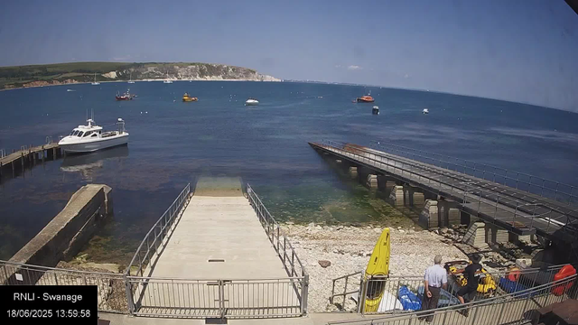 A view of a harbor with several boats floating on the water. There is a white boat docked on the left side near a small pier, and a yellow kayak, along with a few people, are at the bottom right. The background shows a distant shoreline with cliffs and green land under a clear blue sky. A yellow caution sign is visible near the bottom center. The timestamp indicates the date and time as June 18, 2025, at 13:59:58.