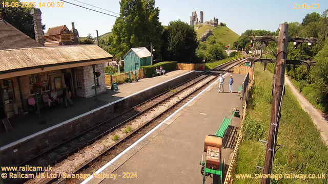 A small train station platform on a sunny day with a few people standing and sitting. There are some benches with people seated, a small green building, and a post with overhead electrical wires for the train. In the background, a hillside with ruins of an old castle is visible, along with green trees lining the station area. The locomotive tracks run alongside the platform, which is bordered by greenery and a dirt path. The timestamp shows the date and time as June 18, 2025, at 14:00:52.