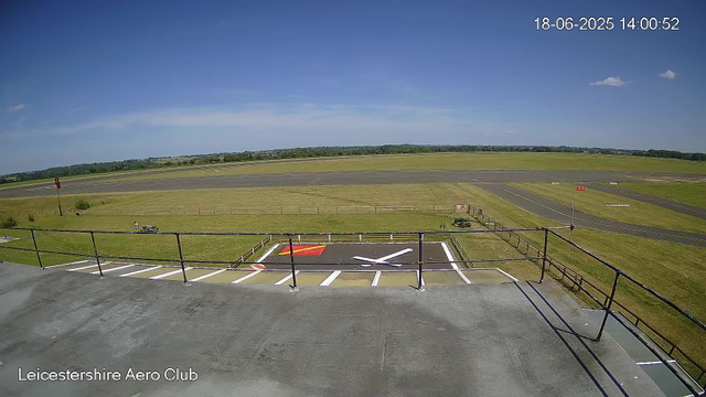 A wide view of an airfield on a clear, sunny day. The foreground shows a terrace with a black railing. There are markings painted on the ground, including a large white "X," a red rectangle, and a yellow arrow. The middle ground features a grassy area with a small white vehicle and a few scattered objects. A runway stretches across the image, with distant trees and open fields beyond. The sky is mostly clear with a few small clouds scattered, and the timestamp in the top right corner reads "18-06-2025 14:00:52." The bottom left corner has the text "Leicestershire Aero Club."