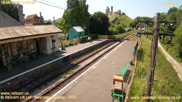 A small train station platform with a stone building on the left, green benches, and flower baskets hanging outside. Three people are standing near a green shed at the far end of the platform, while two others sit on a bench. Overhead power lines run above the tracks, which extend into the distance. On the hill in the background, there are ruins of an old castle. The sky is clear and the scene is brightly lit.
