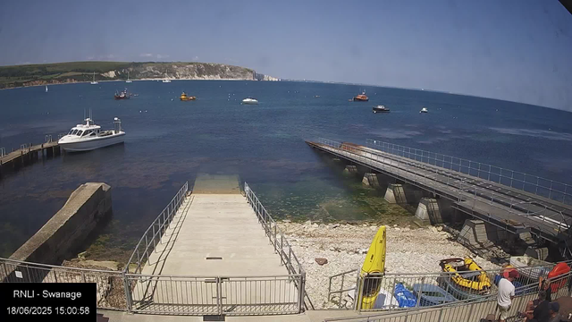 A scenic view of a harbor with several boats and yachts floating on calm blue water. In the background, there are green cliffs and hills under a clear blue sky. In the foreground, there is a concrete dock with metal railings, a small yellow kayak, and a few people standing near the edge. To the right, there is an empty ramp leading into the water and some parked equipment and vehicles. The image has a timestamp and label indicating it was taken at Swannage on June 18, 2025, at 3:00 PM.