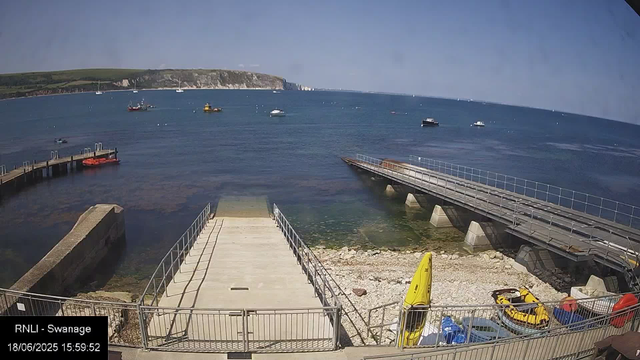 A view of a seaside with boats floating on the water. There are two piers extending into the water, one on the left with some orange objects attached and one on the right with railings. In the foreground, there is a small storage area with yellow and red life-saving equipment, a kayak, and other gear. The background features a green hillside with cliffs meeting the sea under a clear blue sky.