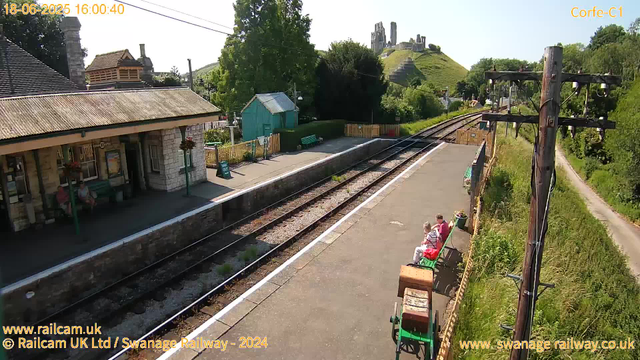 A small train station platform with two people sitting on the benches, one woman in a pink top and the other in a striped shirt, facing each other. There are green benches, potted flowers, and a small shed behind the platform. Railway tracks run alongside the platform, leading towards a hill with ancient ruins atop it in the background. A tall utility pole with wires is on the right side, and a narrow pathway runs parallel to the railway line. The sky is clear and sunny. The top left corner displays the date and time as "18-06-2025 16:00:40".