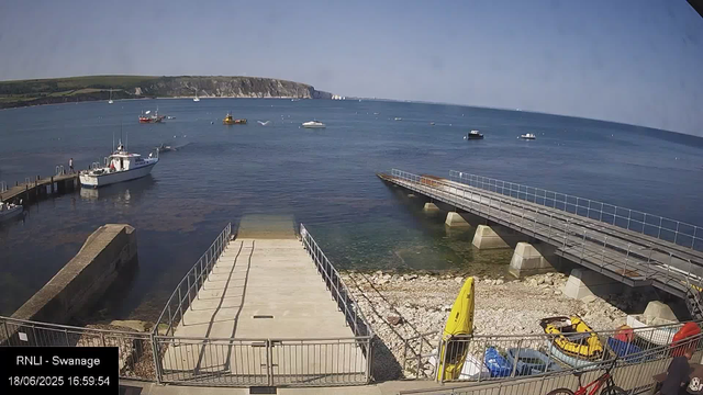 A view of the harbor at Swansea on a clear day, showing a pier with boats docked, calm blue water with several boats and sailboats in the distance, and a coastline with cliffs and green hills in the background. There is also a yellow kayak, a yellow surfboard, and some parked cars along the beach area.