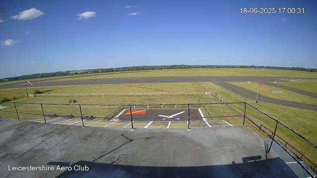 An outdoor view from a webcam shows a clear day at Leicesterhire Aero Club. In the foreground, there is a small helipad with a white helicopter symbol and a red marking. Surrounding the helipad is a wooden and metal fence, with a bench and a flagpole to the left, flying a flag. Beyond the fence, there are taxiways and runways stretching into the distance, bordered by grassy areas. The sky is bright blue with a few scattered white clouds, and the scene is well-lit by sunlight. The date and time stamp at the top right shows 18-06-2025 17:00:31.