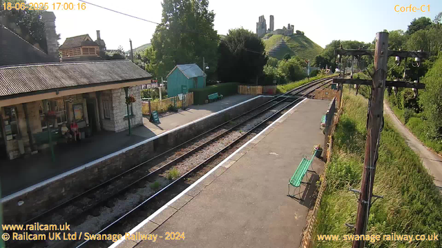 A small railway station platform with a stone building on the left, featuring a bench with two people sitting. There is a signboard on the platform, a bench painted green, and a small teal-colored wooden shed in the background. A pathway runs alongside the platform with a fence separating it from a grassy area with trees. Overhead, there is a utility pole with electrical wires, and on the right, a grassy embankment leads up to a hill with ruins of an old castle or fortress at the top. The sky is clear and sunny. The image is marked with the date and time: 18-06-2025, 17:00:16.