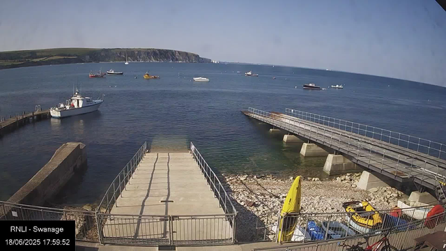 A view of a harbor with boats anchored on calm water. In the foreground, there is a concrete pier with metal railings, and a small yellow kayak is resting on the rocks nearby. To the right, there is a yellow lifeguard station and some small boats and equipment. In the background, there are several boats scattered across the water, and a cliff or headland covered with greenery extends into the sea on the horizon. The sky is clear with some light clouds.