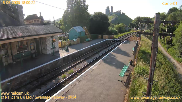 A small railway station platform with a building on the left side featuring a slate roof and stone walls. There are flower baskets hanging by the building entrance. In the middle distance, there is a small green shed with a pitched roof. The railway tracks run parallel to the platform, curving toward the right, with overhead electric lines supported by wooden poles on the right side. Several benches are on the platform, and a sign indicating "Way Out" is visible near the building. In the background, a lush hillside with trees and what appears to be old ruins or structures on top is visible, bathed in sunlight.