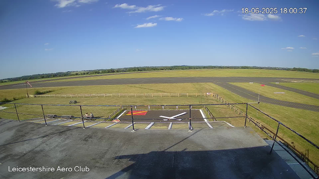 A view from a webcam showing the Leicesterhire Aero Club. The scene includes a small parking area in the foreground with designated spaces and a helipad marked with a large white "X" and red and yellow markings. Beyond the parking area, there is a grassy field enclosed by a wooden fence, with a bench and two people sitting on the grass. The background features a runway stretching across the landscape, with a clear blue sky, a few scattered clouds, and distant trees on the horizon. The timestamp in the top right corner indicates it is June 18, 2025, at 6:00 PM.