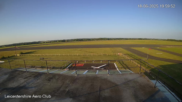 An empty rooftop helipad at Leicesterhire Aero Club, with a small red and white circle marking the landing area. A British flag is flying on a flagpole, and there are wooden fences around the helipad. Beyond the helipad, there is a grassy area, a runway, and open fields under a clear blue sky. The top right corner displays the date and time: 18-06-2025 19:59:52.