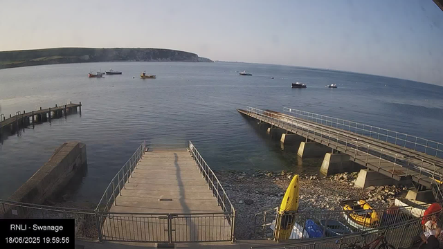 A view of a seaside dock with calm water and boats anchored offshore. There are two piers extending into the water, one on the left and one on the right, with a rocky shore below. In the foreground, there are yellow safety equipment and a person with a red backpack near the fence. In the background, a headland stretches across the horizon under a clear sky.