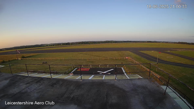 The image shows a tarmac area with a large clock painted on the ground, positioned lower center of the image. Behind it is a grassy area enclosed by a fence, with a small flagpole featuring a waving flag on the left side. In the background, there are runways and open fields extending into the horizon, and a clear sky with the sun setting, casting a warm glow. The timestamp in the top right corner reads "18-06-2025 21:00:33." In the bottom left corner, text indicates "Leicestershire Aero Club."
