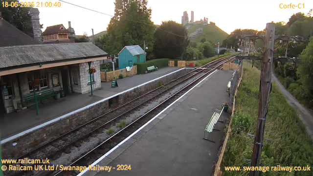 A small train station platform at sunset, with two green benches and a green building. Railway tracks run in front of the platform, and there are two wooden benches along the edge. Power lines and a willow tree are visible to the right, with a hill and a castle ruins in the background. The image is time-stamped "18-06-2025 21:00:40."
