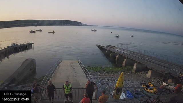 A view of a waterfront at sunset shows a small group of people gathered near a railing in the foreground, with some wearing high-visibility vests. There is a concrete ramp leading into the water, and several boats are anchored in the calm sea. In the background, the horizon features distant cliffs or headlands, and the sky is clear with soft pastel colors. A few green algae or moss patches are visible on the rocky shoreline below the ramp.