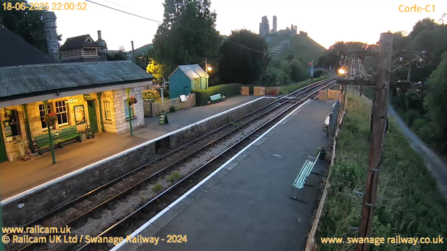 A small train station platform is illuminated by outdoor lights during dusk, with a station building featuring a green door and windows decorated with flower baskets. Adjacent to the building, there are two benches and a "Way Out" sign. A railway track runs parallel to the platform, with a wooden fence separating the platform from a grassy area on the right. In the background, a hillside with a castle or ruins is silhouetted against the evening sky, and electrical poles with wires run along the right side of the scene. The timestamp shows the date and time as June 18, 2025, at 10:00:52 PM.