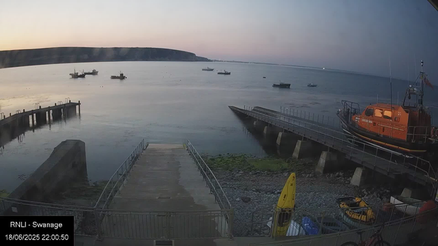 A view of a calm harbor at dusk with several small boats floating on the water, a concrete pier extending into the bay, and a bright orange rescue boat docked on a pier to the right. The sky is tinged with soft pastel colors from the setting sun, and distant cliffs line the horizon. A yellow kayak is positioned on the rocky shore near the bottom right corner. The scene appears peaceful and quiet.