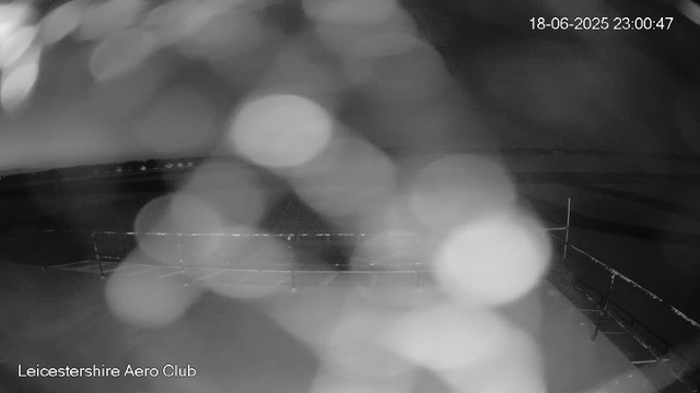 A black-and-white image from a webcam shows a nighttime scene over a body of water at Leicestershre Aero Club. The image is blurry with large, out-of-focus light spots. A distant shoreline is visible with some faint lights, and a sailboat with a visible mast is on the water near the bottom right. The timestamp in the upper right corner reads 18-06-2025 23:00:47.