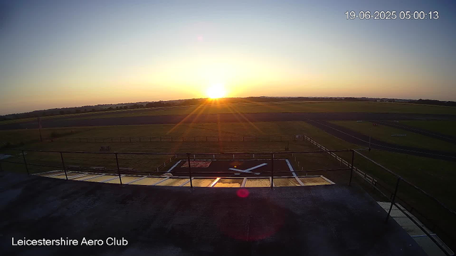 The image shows a runway and a helicopter pad at the Leicestershire Aero Club during sunrise, with the sun on the horizon casting a warm glow across the fields and sky. The timestamp indicates it was taken on June 19, 2025, at 5:00 AM. The scene is viewed from an elevated position, possibly a balcony or observation deck.