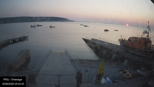 A sunrise over a harbor with several boats floating on calm water. A red rescue boat is docked on the right, and a pier extends into the water on the left. A person is standing near the lower center of the image, looking towards the harbor. The sky is beginning to brighten with soft pastel colors.