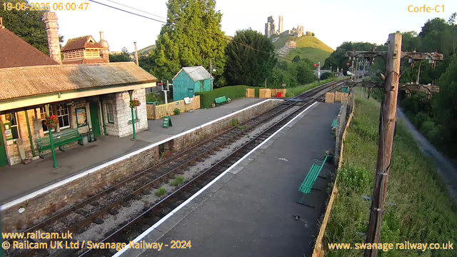 A small train station platform with a ticket office building on the left, decorated with hanging flower baskets. The platform has green benches and a "Way Out" sign. There is a small green shed and a wooden fence with plants and a clothesline on the far side. The railway tracks run down the middle, with a grassy area and trees to the right. In the background, a hill with a castle ruins at the top is visible. An orange utility pole with wires stands on the right side near the edge of the platform. The sky is clear and bright, indicating early morning. The image has labels indicating it was taken on June 19, 2025, at 6:00 am.
