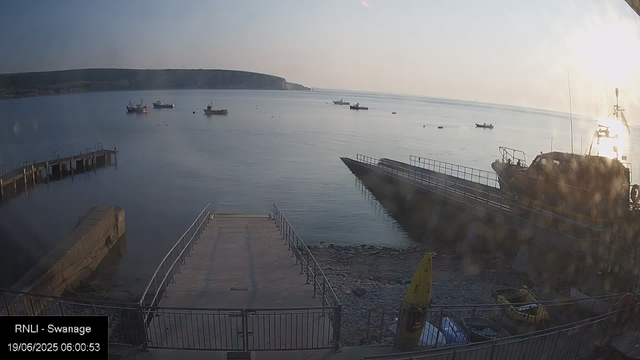 A calm harbor scene on a clear morning with boats anchored on still water, a small pier on the left, a large docked boat on the right, and a hilly shoreline in the distance under a bright sky.