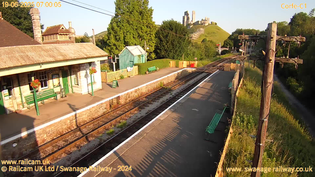 A quiet local railway station platform bathed in warm morning sunlight. The platform is lined with green benches and hanging flower baskets. There are a few small structures, including a green shed and a ticket office, with a "Way Out" sign near the entrance. Overhead, electrical wires run along wooden poles. In the background, a green hillside is topped by a historic castle or fortress ruin perched on a hill. The scene is framed by trees and lush greenery on either side.