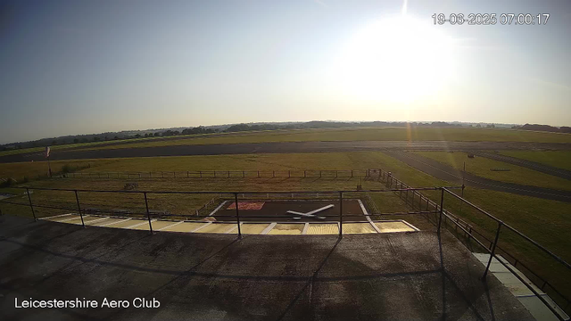 A clear morning scene at LeicesterShire Aero Club, with the sun rising over a flat landscape of green fields and open sky. The foreground shows part of the rooftop and a yellow wind sock on a pole to the left. The times are displayed in the upper right corner, indicating early morning hours.