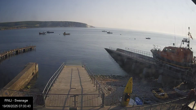 A tranquil harbor scene with a concrete pier extending into calm water, a bright orange rescue boat moored on the right side, several boats floating in the distance, a yellow kayak near the bottom right, and a rocky shoreline in the foreground. The sky is clear with soft lighting, and distant cliffs stretch along the horizon.