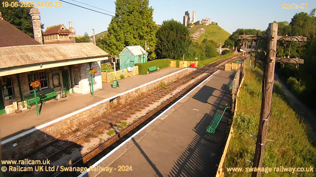A small countryside train station platform bathed in morning sunlight, with green benches and flower planters near the waiting area. Railway tracks run through the scene, with a small green shed in the background, surrounded by lush trees and hills. In the distance, a castle sits atop a hill, overlooking the landscape. Overhead, electrical wires stretch across the sky, and a tall wooden utility pole is on the right side. The timestamp on the image indicates it was taken on June 19, 2025, at 8:00 AM.