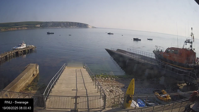 A calm harbor scene with several boats anchored in the water. There is a dock on the left with a white boat tied to it. A larger red rescue boat is on the right, partially on a ramp. In the foreground, there is a small boat and yellow buoy. The background features a distant shoreline with white cliffs and green hills under a clear sky. The image is timestamped June 19, 2025, at 8:00 AM in Swansea.