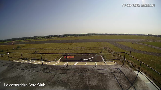 A wide view of an airfield from a webcam, showing a clear sky with the time and date stamp "19-06-2025 09:00:54" in the top right corner. There is a helicopter landing pad in the foreground with a helicopter's tail rotor visible, surrounded by a black fence. In the background, open grassy areas and runways extend into the distance under bright sunlight. The bottom left corner has the text "Leicestershire Aero Club."