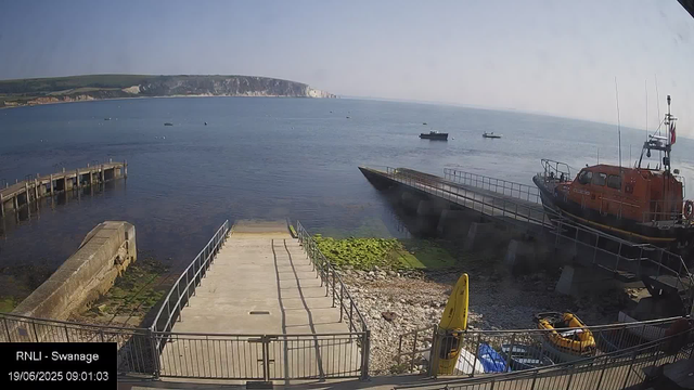 A view of a harbor with a concrete pier and a boat ramp leading into the water in the foreground. On the right side, an orange rescue boat is parked on a slipway, with a yellow inflatable rescue craft nearby. Several small boats are floating on the calm water, and a large cliff with white chalk or limestone cliffs stretching along the coastline is visible in the background. The sky is clear with a few light clouds, and the overall scene appears to be in the morning.