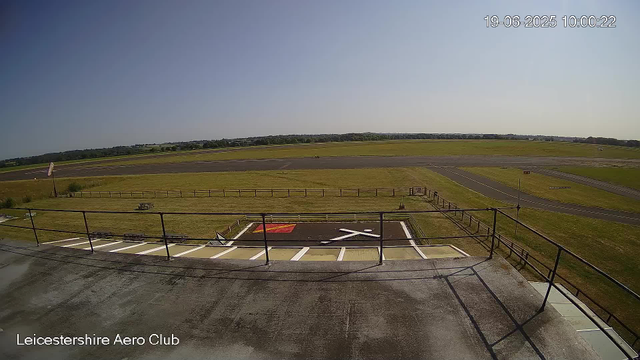 A view from a webcam at Leicestershire Aero Club showing an empty parking area with designated spots, a small grassy area with a bench and a flagpole, and a runway extending into the distance under a clear blue sky with a few distant trees on the horizon. The timestamp in the top right corner indicates it was taken on June 19, 2025, at 10:00:22.