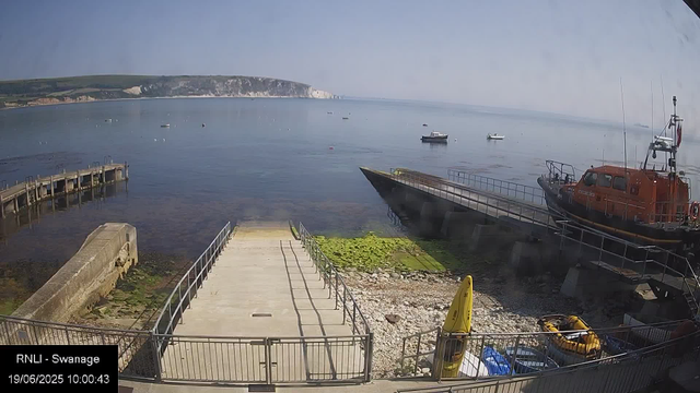 A view of a calm seaside with a dock, boats floating in the water, and a rugged shoreline in the background. There is a large orange rescue boat on the right side, a small yellow inflatable boat near the bottom, and some equipment and barriers along the waterfront. The sky is clear with some light haze over the horizon. The timestamp indicates the image was taken on June 19, 2025, at 10:00 AM.