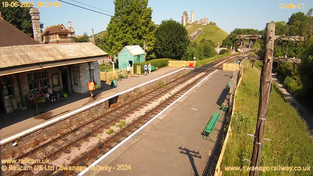 A small train station platform with a stone building on the left, where three people are sitting and one person in an orange safety vest is walking. Two people are walking along the platform. There are green benches and a small green shed in the background. Railway tracks run through the center of the image, with a yellow fence at the far end. A large hill with ruins of an ancient castle atop it is visible in the background, surrounded by lush green trees. A wooden utility pole with wires is on the right side, and a narrow street runs parallel to the tracks on the far right. The sky is clear and sunny. The timestamp indicates the date as June 19, 2025, and the time as 10:01:05.